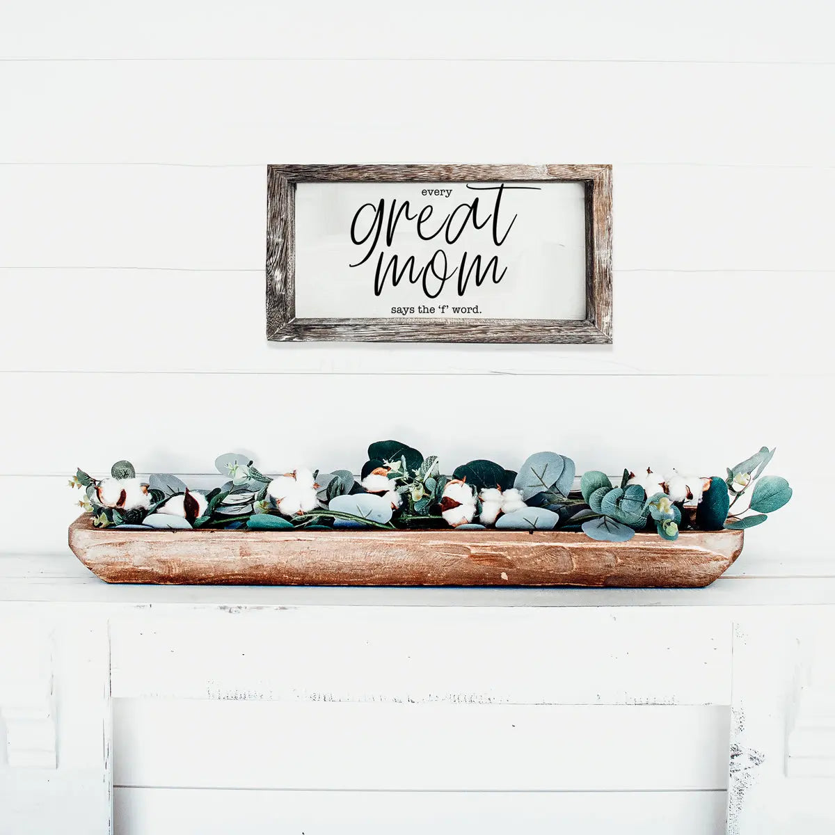Decorative sign with 'every great mom says the f word' text above a wooden tray with greenery on a white surface.