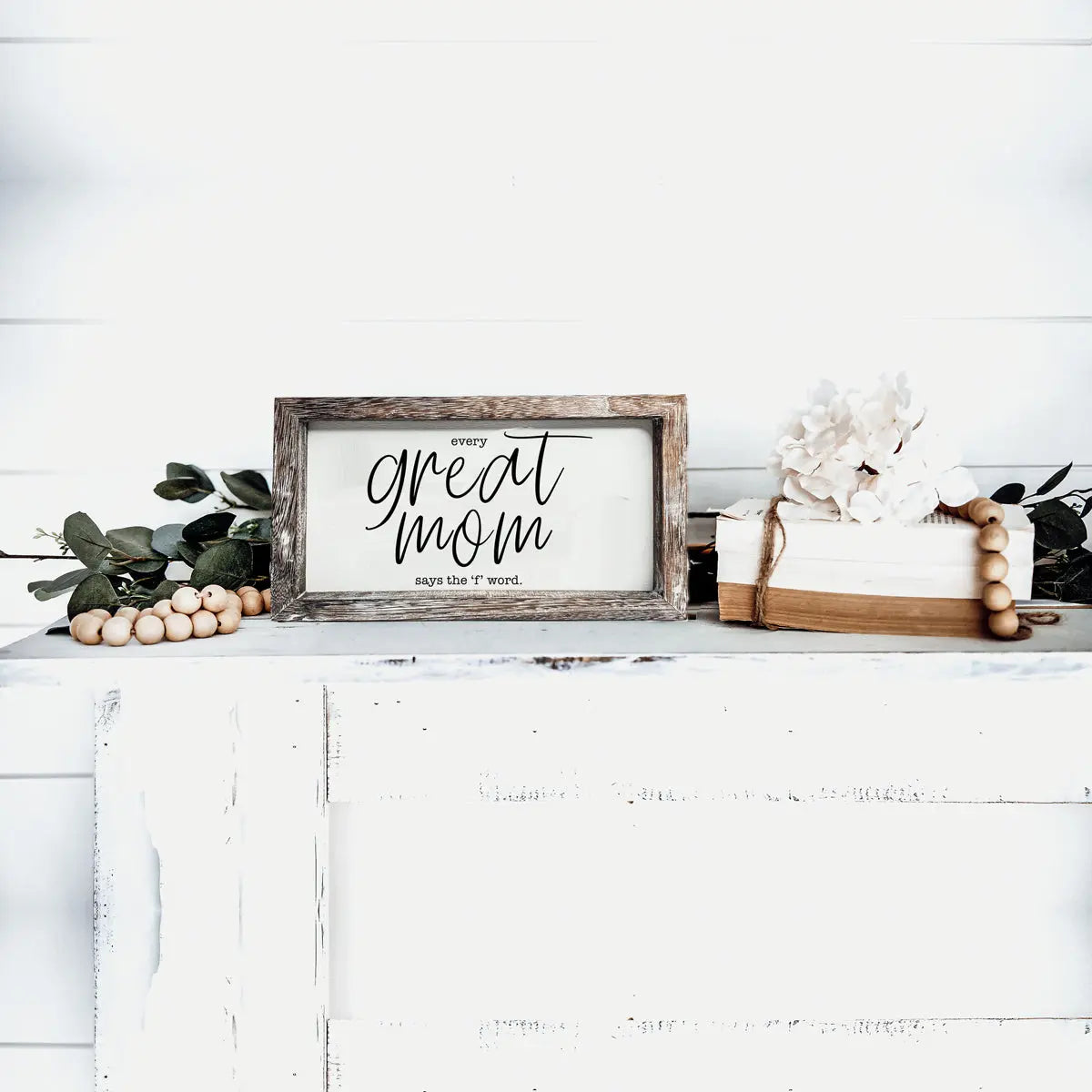 Decorative sign on a white surface with text 'Every great mom says the T word' surrounded by decorative items.