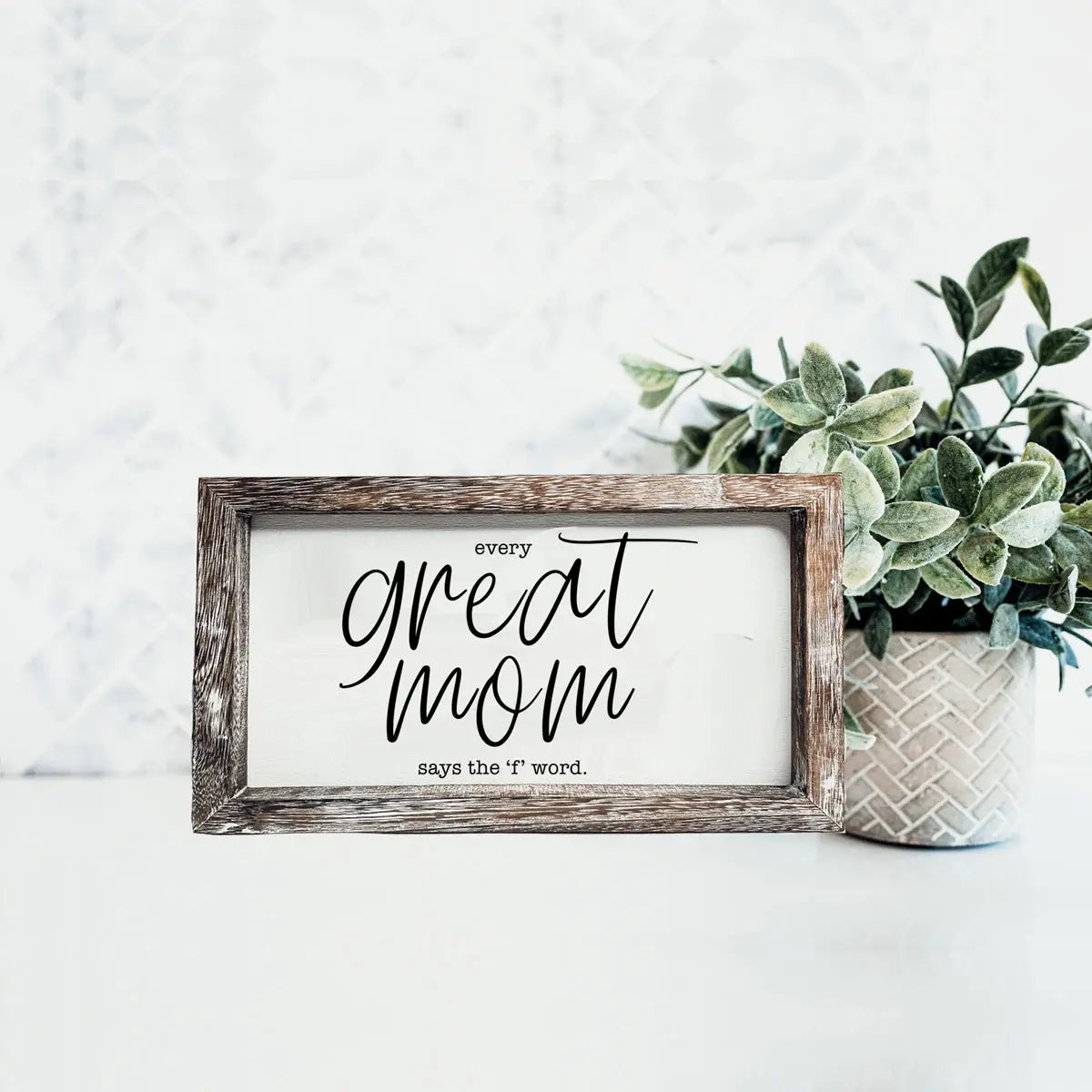 Framed print with text 'Every great mom says the f word' on a white surface with a plant.