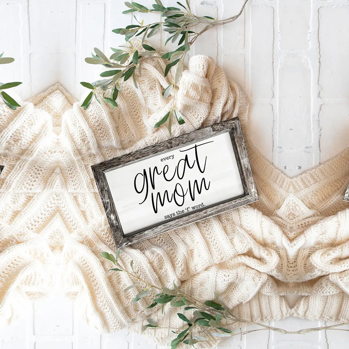 Decorative sign with 'every great mom says the word' on a textured surface with greenery
