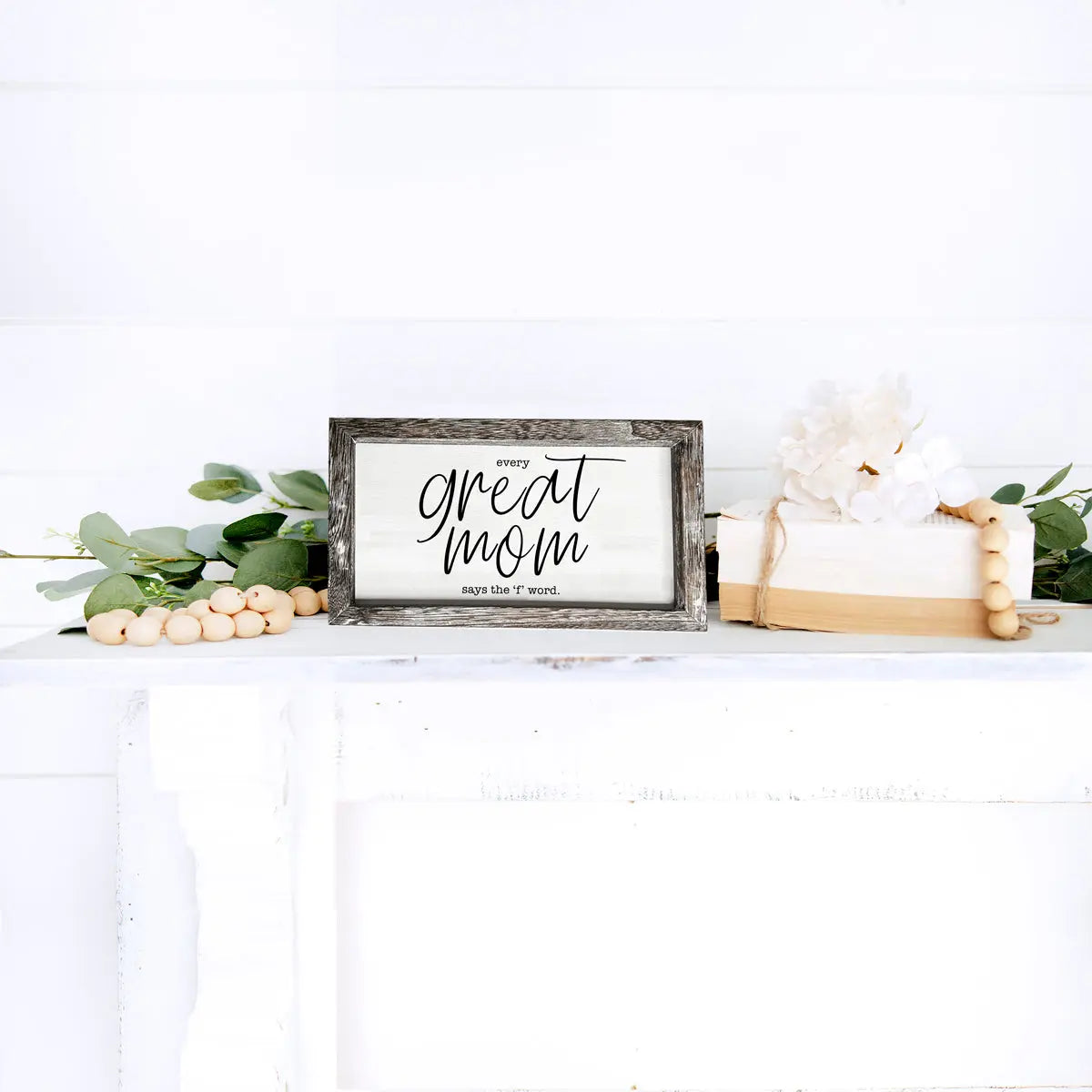 Decorative sign with 'Great Mom says the f-word' text on a white surface with greenery.