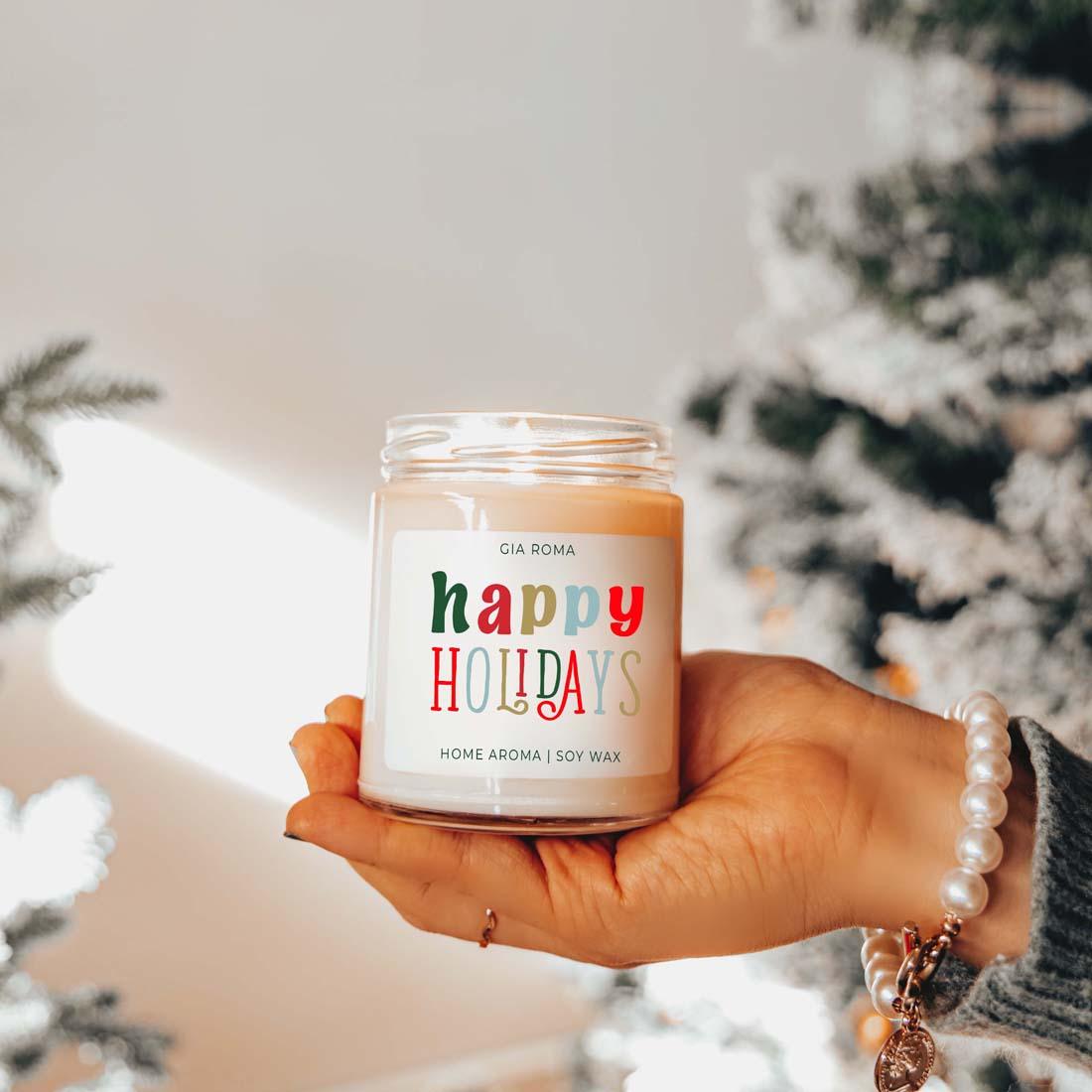 Hand holding a 'happy holidays' candle with Christmas trees in the background