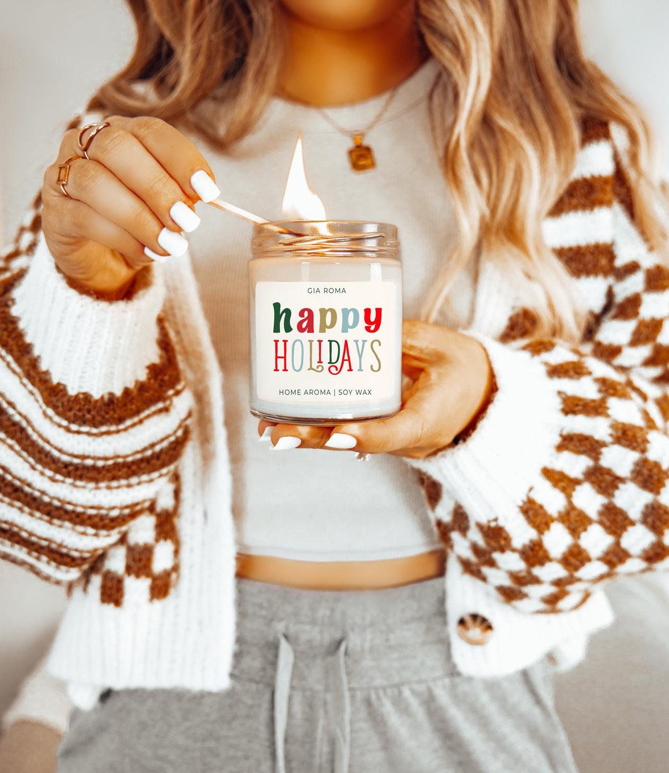 Person lighting a candle labeled 'Happy Holidays' with a cozy sweater and necklace.