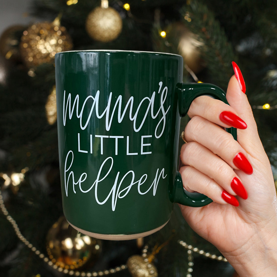 Hand holding a green mug with 'mama's little helper' text in front of a decorated Christmas tree.