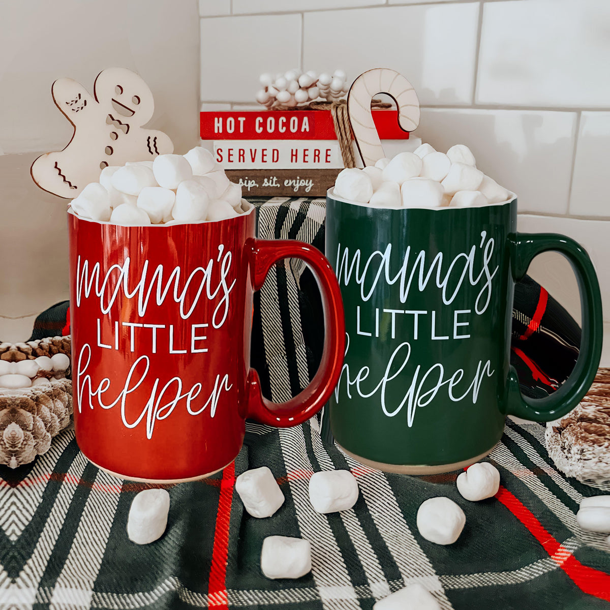 Two mugs labeled 'Mama's Little Helper' with marshmallows on a checkered tablecloth.