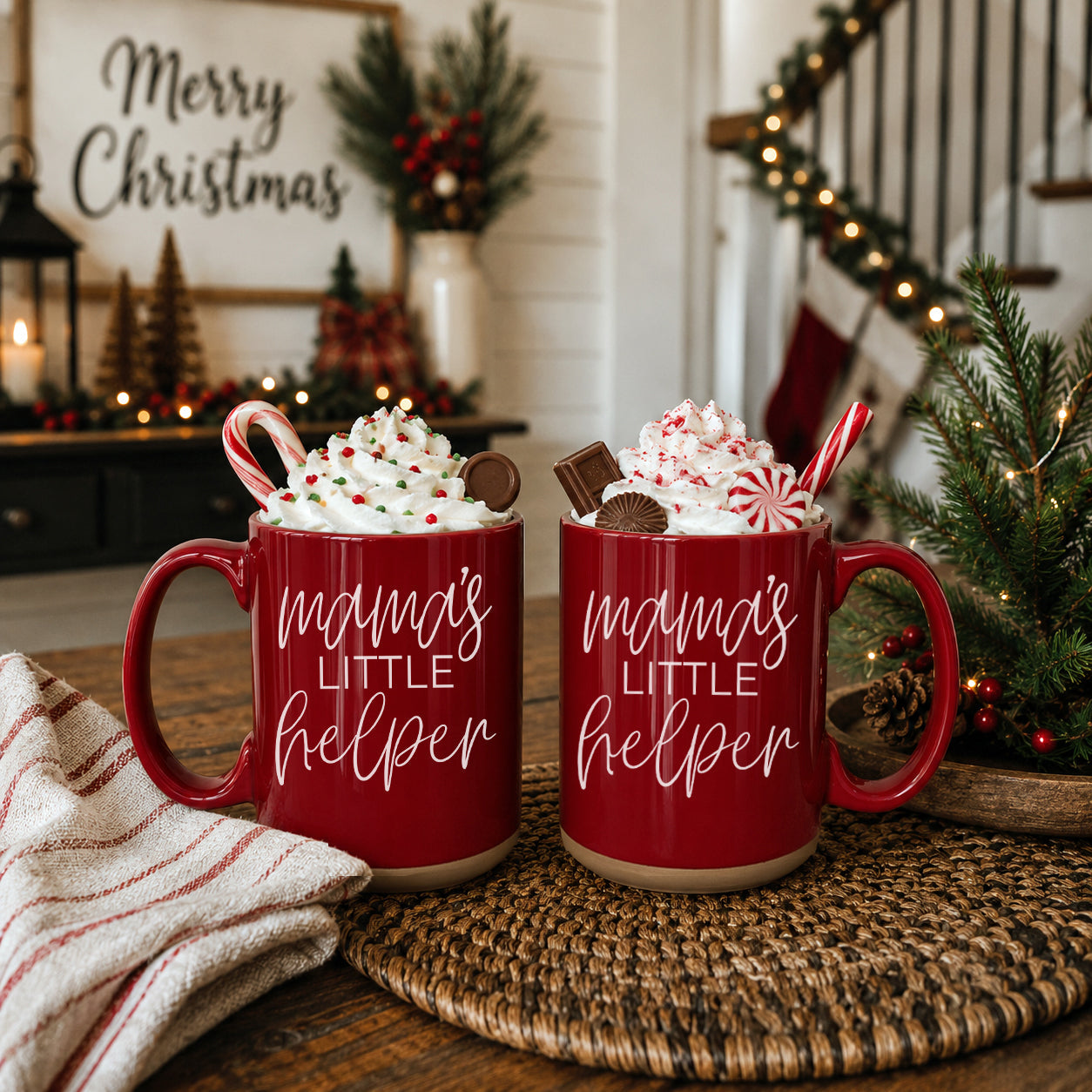 Two red mugs with 'mama's little helper' text, filled with hot chocolate and candy canes, on a table with Christmas decorations.