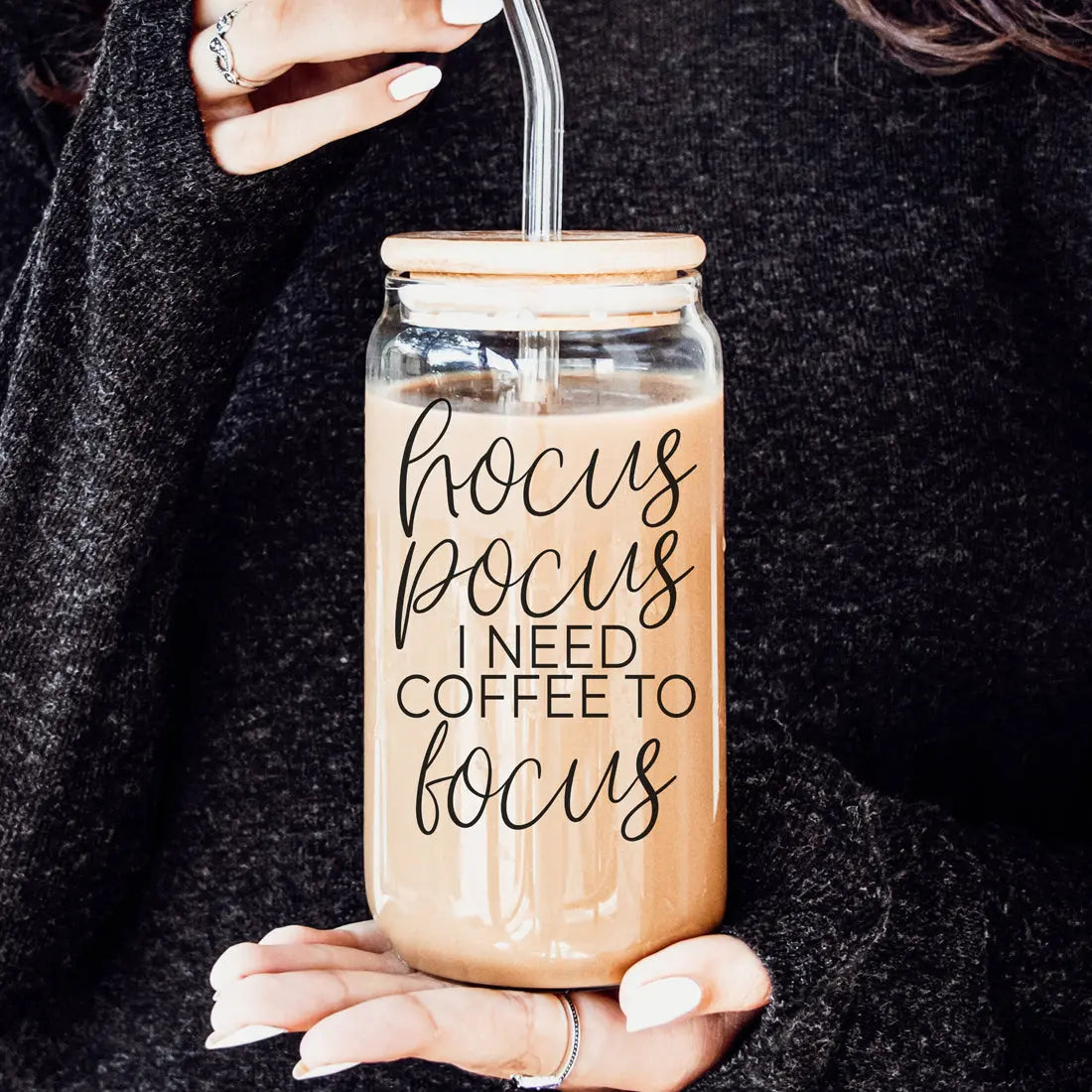 Glass jar with straw lid filled with coffee, featuring text 'hocus pocus I need coffee to focus', held by a person.