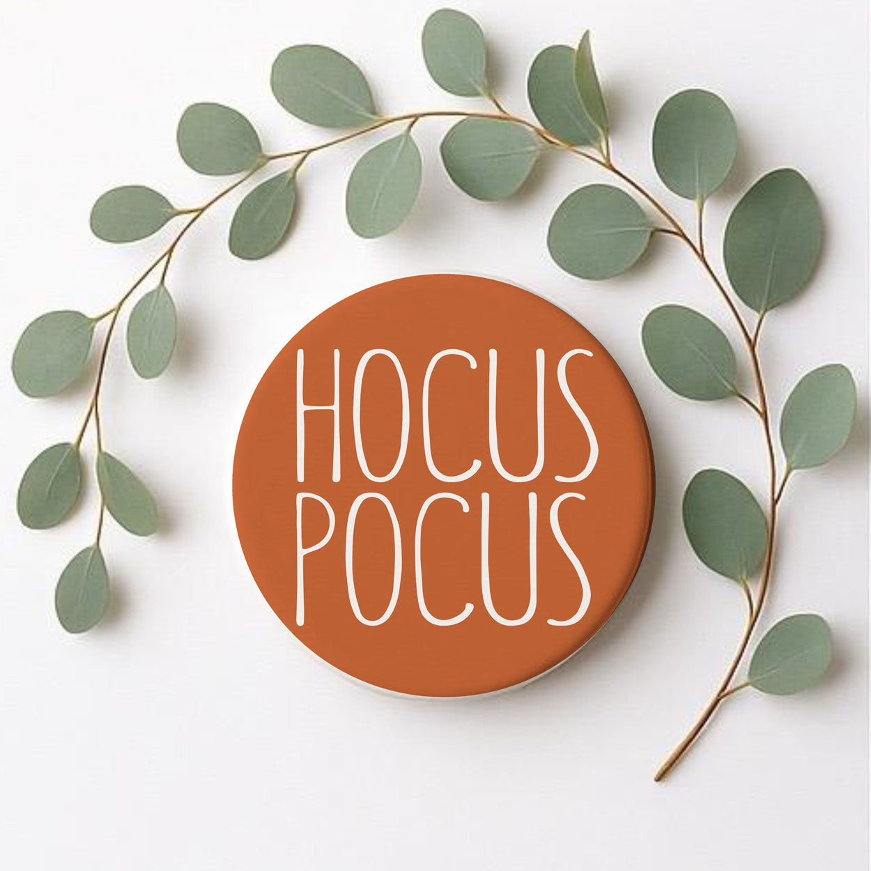 Round sign with 'HOCUS POCUS' text surrounded by green leaves on a white background