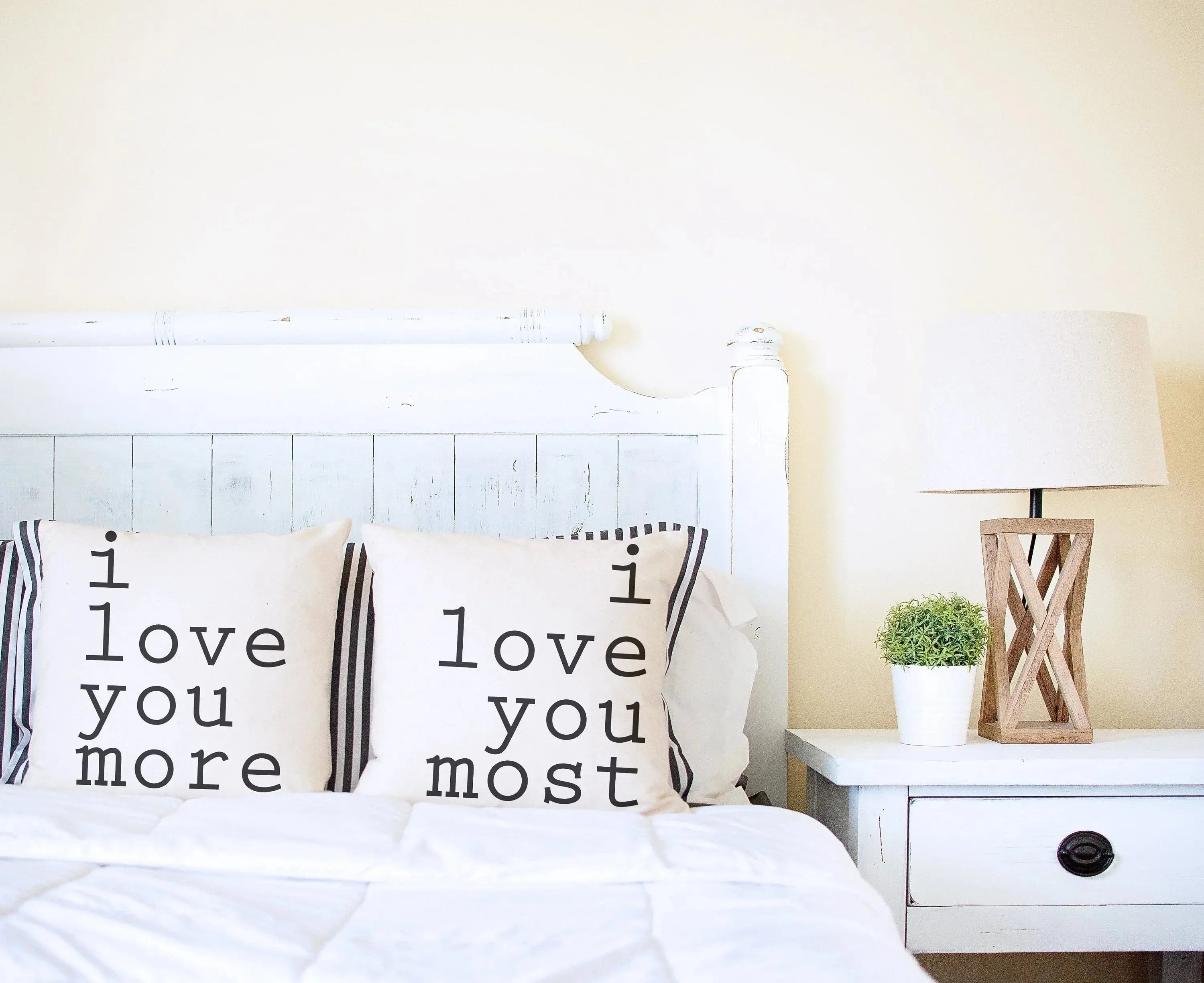 Cozy farmhouse bedroom with 'I love you more' and 'I love you most' throw pillows on white bed, nightstand, lamp, and plant