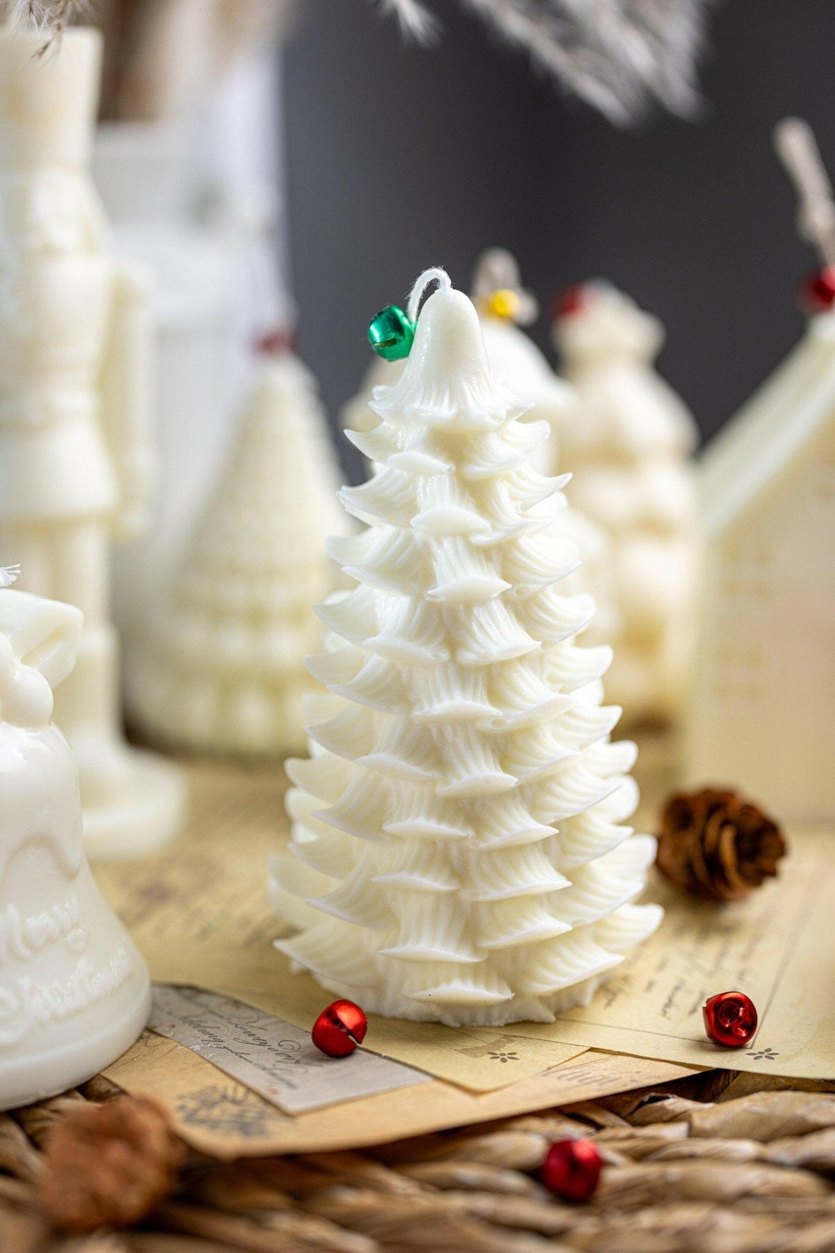 A white Christmas tree-shaped candle with layered textured branches, decorated with small red and green jingle bells, displayed on a rustic surface with other holiday candles in the background.