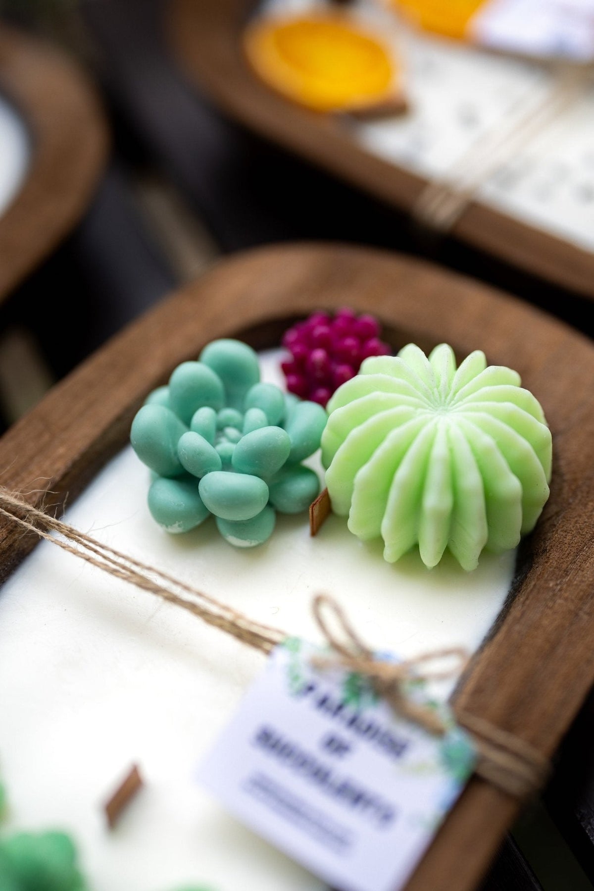A close-up of a wooden tray holding a white candle with decorative sculpted soaps in mint green and pale yellow, plus a small purple soap and a paper tag tied with twine.