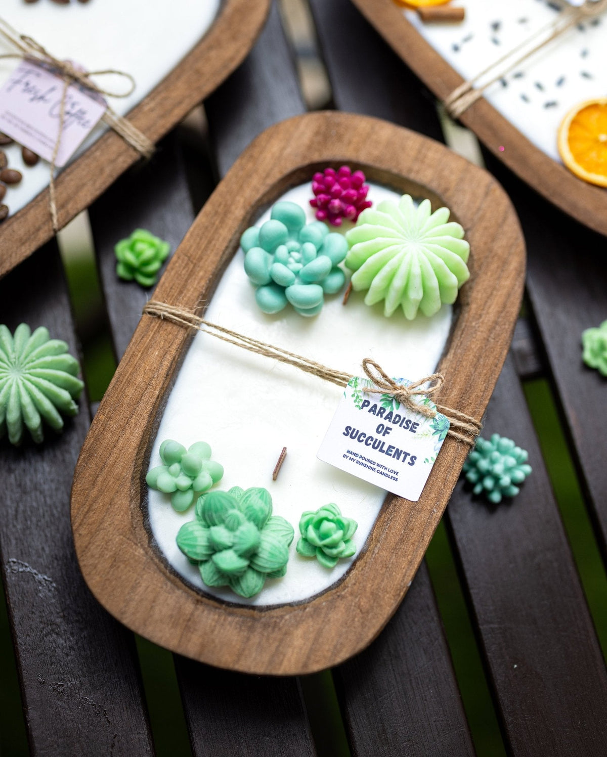 A close-up of a succulent-themed candle in an oval wooden tray with a white wax base and raised green succulent designs, tied with twine and a small tag reading 'Paradise of Succulents.'.