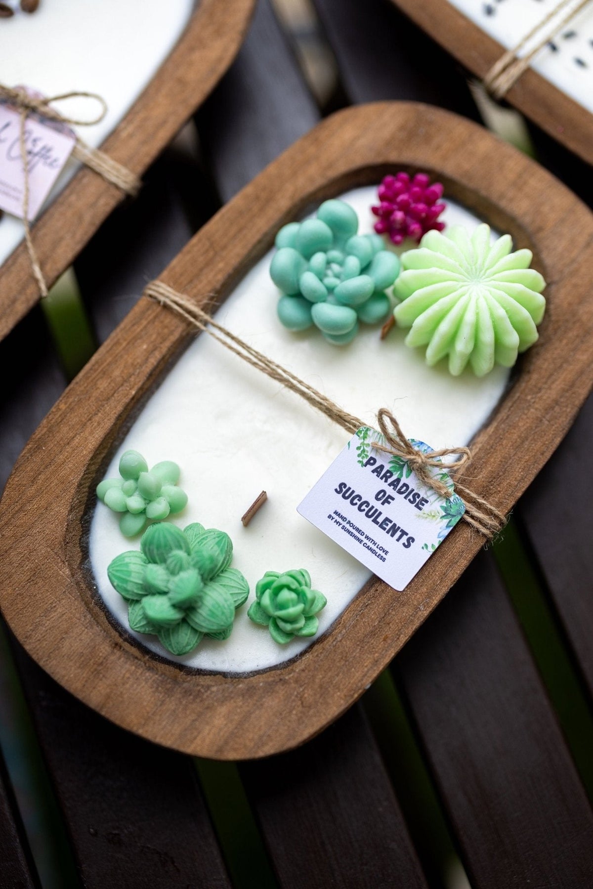 A close-up of a wooden tray containing a white wax melt or soap base decorated with sculpted succulent shapes in shades of green, mint, and magenta. A small paper tag is tied to the center with twine.