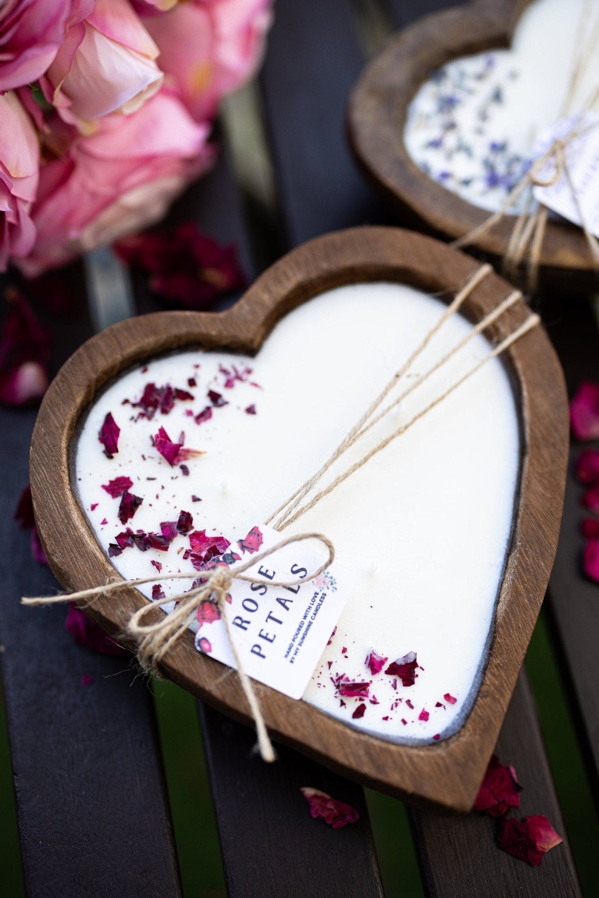 Heart-shaped wooden candle holder containing a white candle with dried rose petals on top, tied with twine and a tag reading ROSE PETALS, on a dark wooden surface with pink flowers in the background.