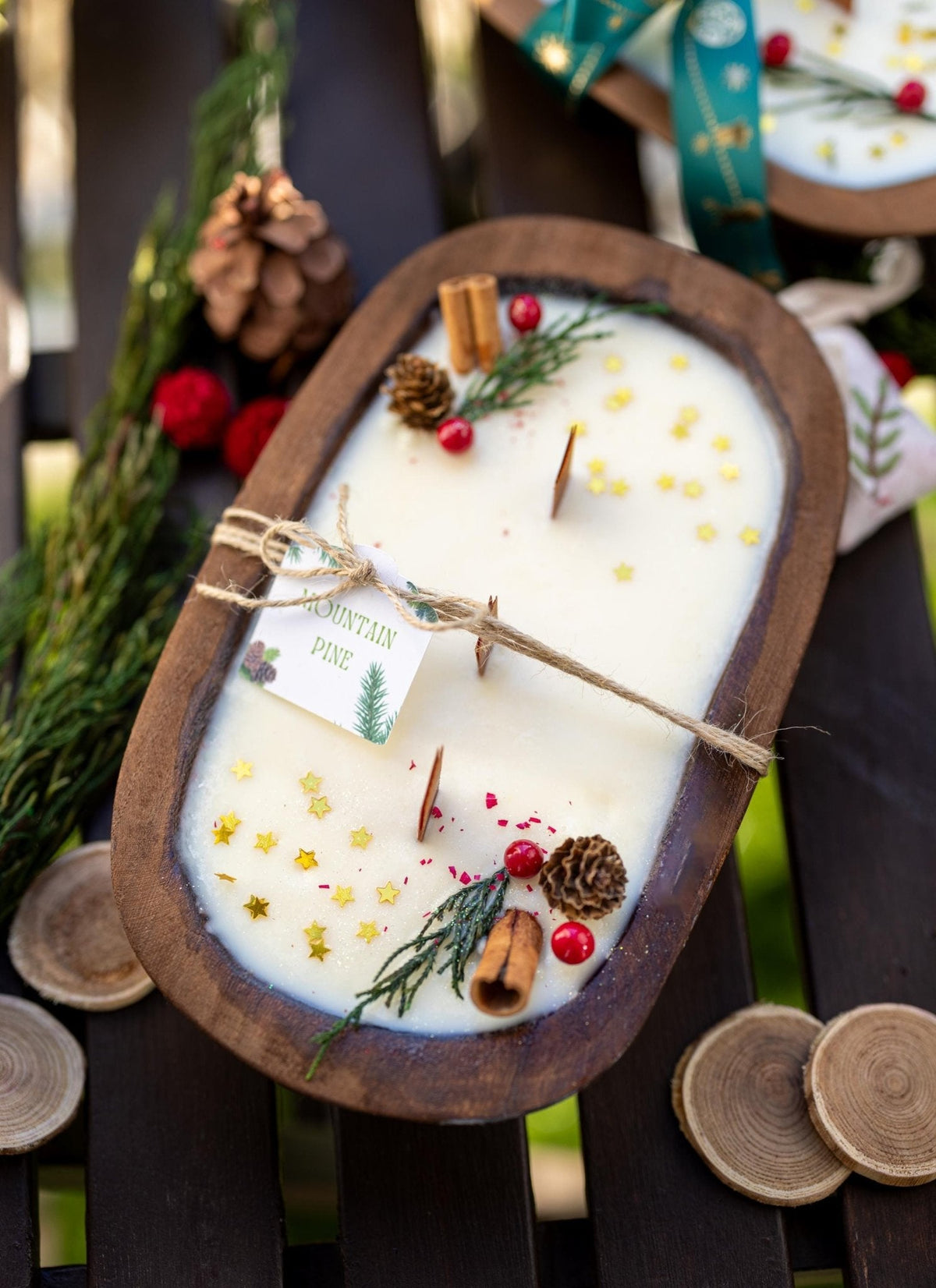 An oval wooden candle holder filled with white wax and decorated with pinecones, cinnamon sticks, red berries, and evergreen sprigs, tied with a twine string and a small tag that reads 'Savoryn Pine.' It is styled on a dark wooden slatted surface with holiday greenery and wood slices in the background.