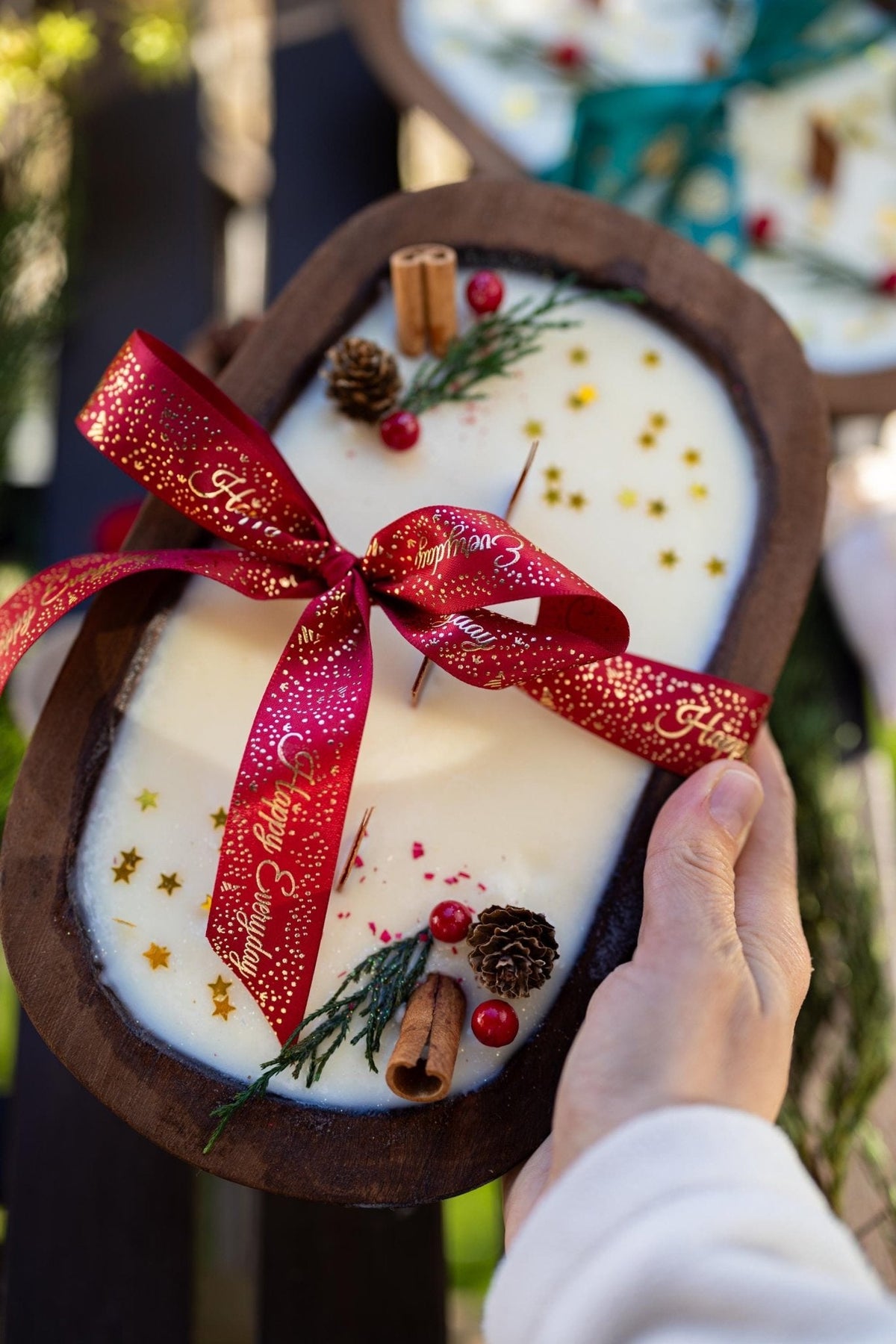 Decorative candle with a red ribbon, cinnamon sticks, and pine cones in a hand.
