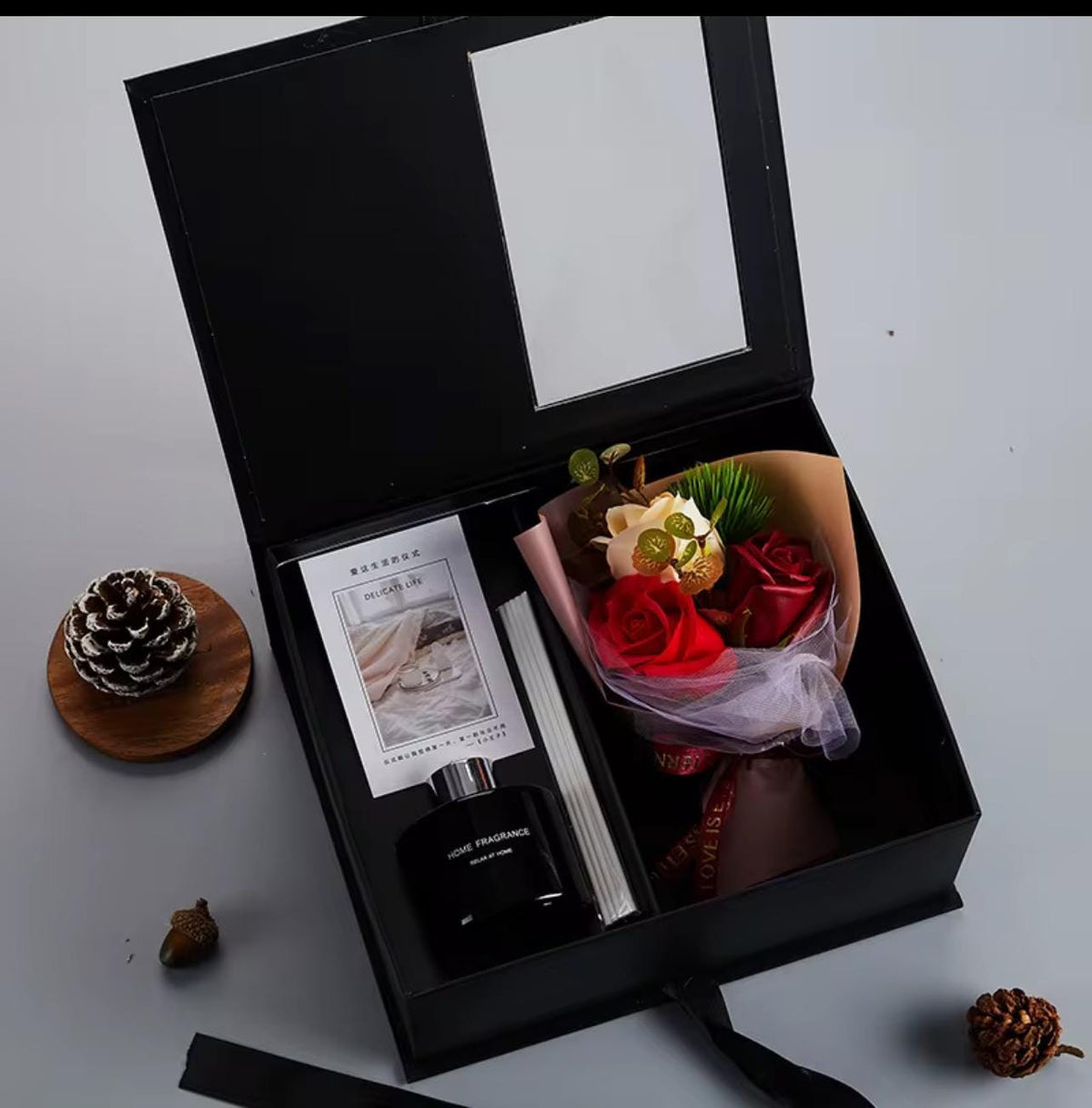An open black gift box containing a black perfume bottle, a printed card, and a wrapped bouquet of red and cream flowers, arranged on a light gray surface with small decorative pinecones around it.