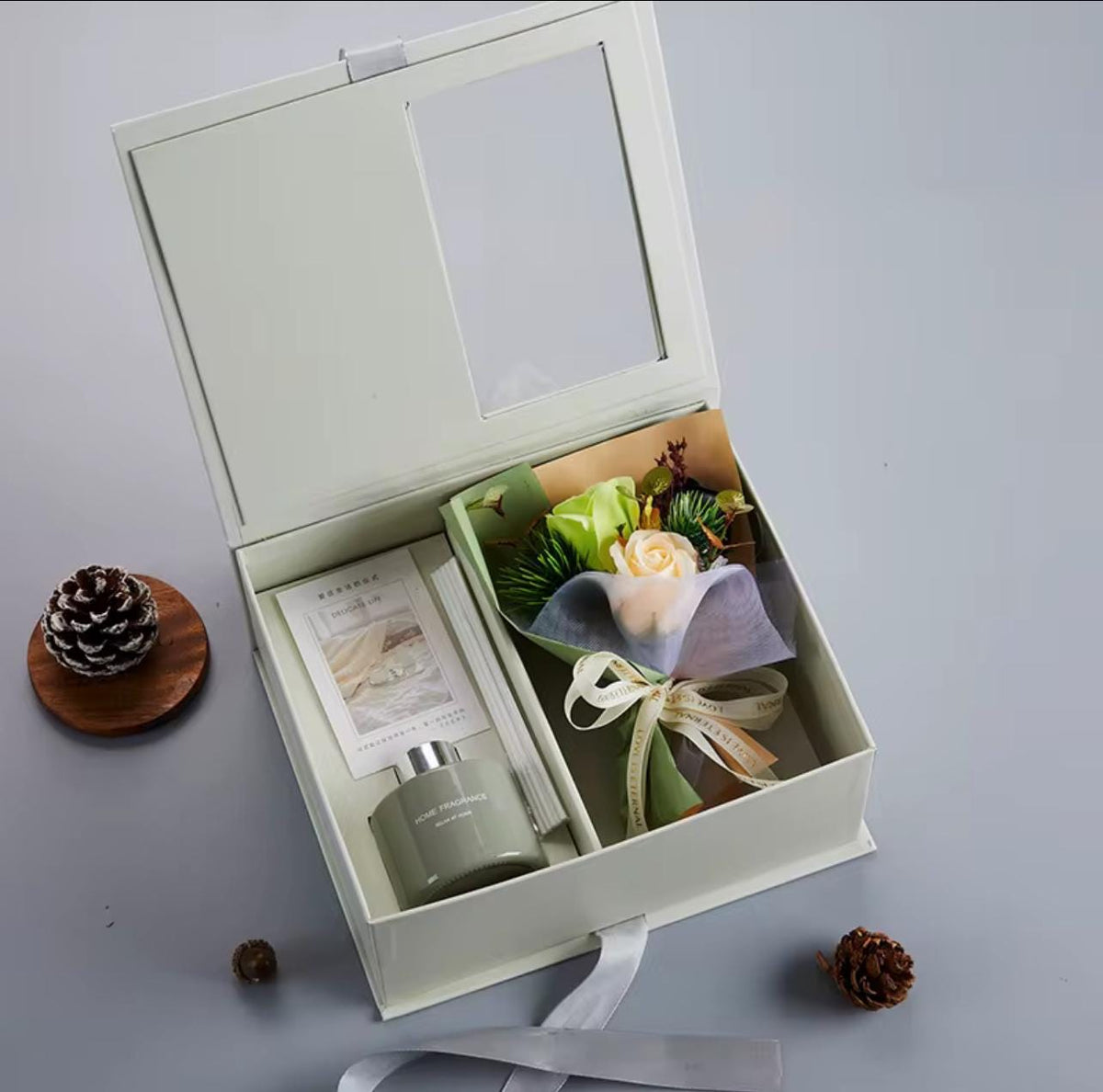 An open cream-colored gift box with a clear window lid, containing a small green candle, a card, and a mini flower bouquet. The box is styled on a light gray surface with decorative pinecones.