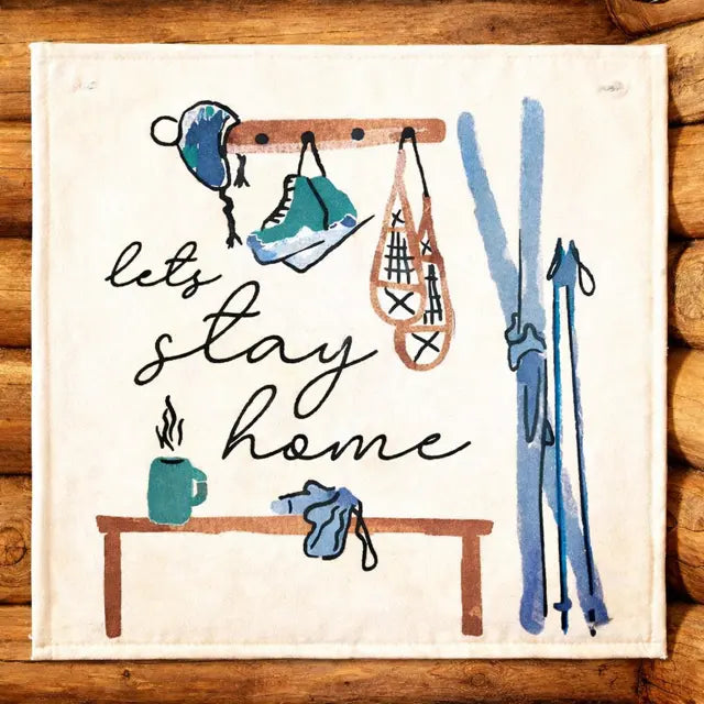 A square cream-colored fabric napkin or tea towel featuring a rustic winter sports illustration with the phrase 'let's stay home' in black script. The design includes blue and teal skis, a blue beanie, a teal shoe, orange-brown skates, and a small teal mug on a wooden table, all set against a background of stacked wood logs.
