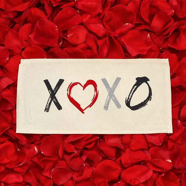 A rectangular cream-colored fabric napkin or coaster printed with the letters 'XOXO' in black, red, and gray brushstrokes, laid flat on a dense bed of red rose petals.