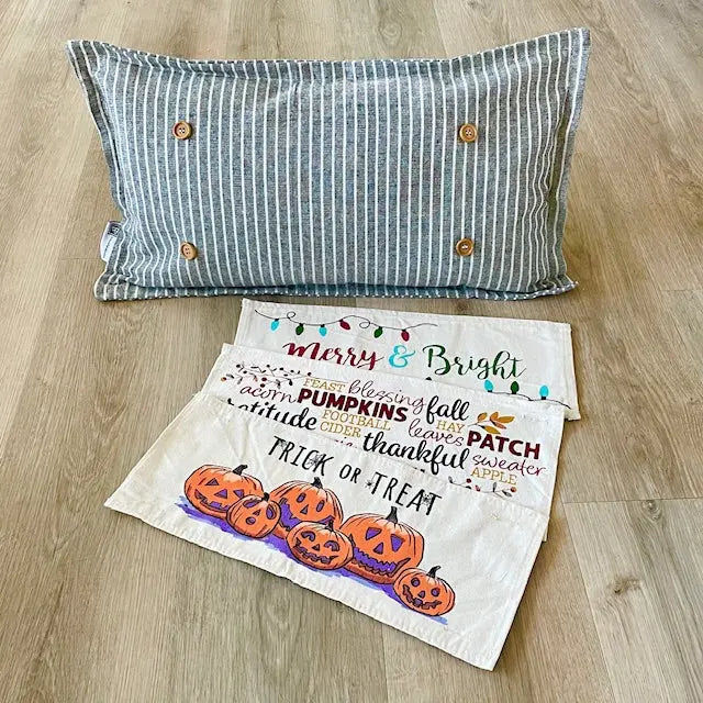 A striped blue and white pillowcase with wooden button details is displayed on a light wood floor next to three white fabric napkins featuring a colorful autumn 'Trick or Treat' word cloud and pumpkin illustrations.