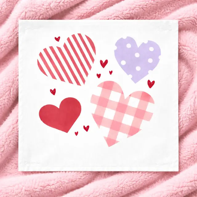 A square white fabric napkin or coaster featuring a heart pattern with red stripes, a red polka dot heart, a red solid heart, and a pink gingham heart, displayed on a pink plush blanket.