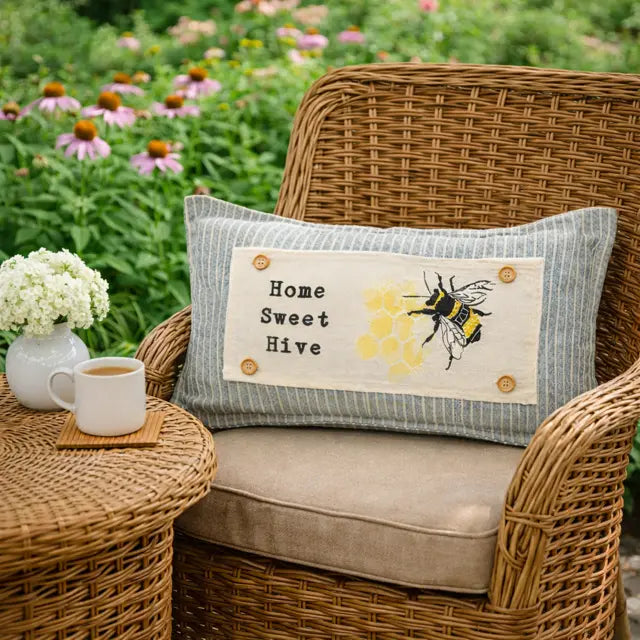 A rectangular decorative throw pillow with a striped blue and white border and a central cream panel reading 'Home Sweet Hive' with a bee and honeycomb design, resting on a wicker patio chair in a garden setting.