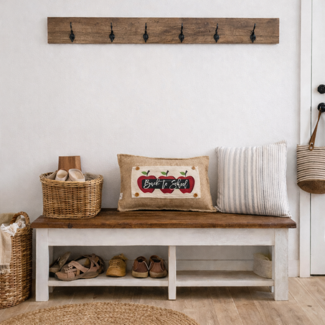 A rustic white bench with a dark wood seat in a bright entryway, styled with a burlap pillow that says 'Back to School' with red apples, a striped pillow, shoes in the shelf, and a wicker basket.