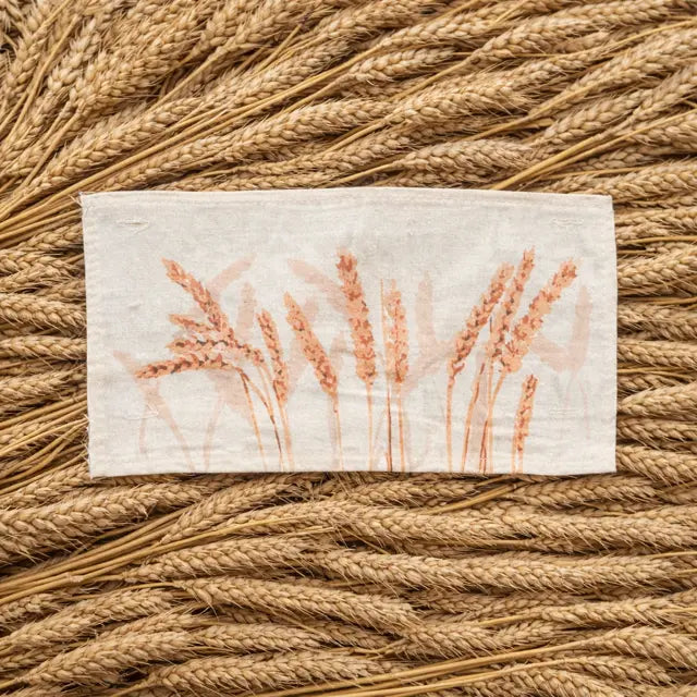 A square off-white linen cloth featuring a printed design of wheat stalks in a warm terracotta color, laid flat on a textured background of dried wheat stalks.