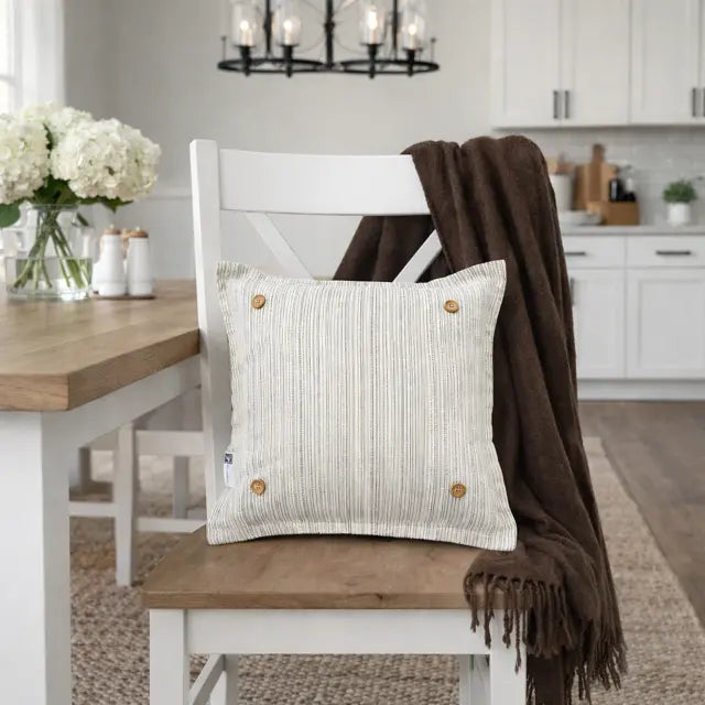 A square off-white ribbed throw pillow with four wooden buttons, styled on a white wooden dining chair with a brown throw blanket draped over it, set in a bright, modern kitchen.
