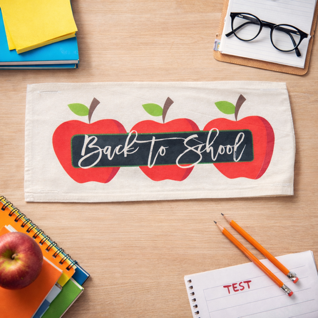 A rectangular cream fabric item printed with three red apples and a black chalkboard-style banner reading 'Back to School' in white script, shown on a wooden desk with notebooks, pencils, glasses, and an apple around it.