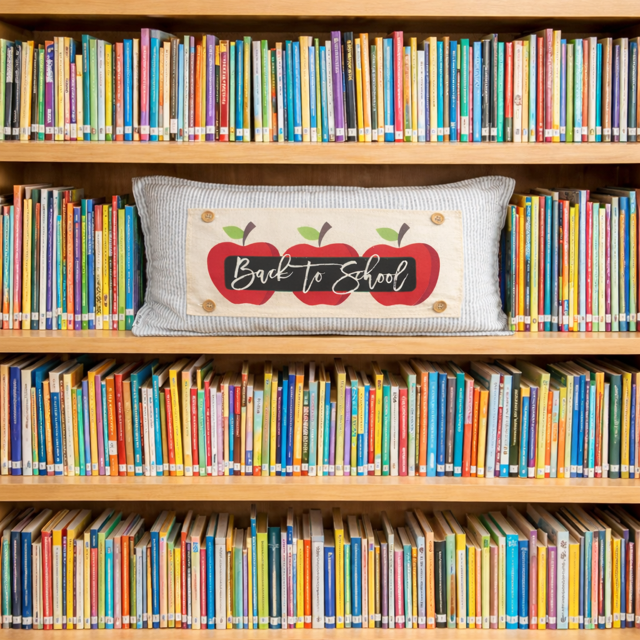 A rectangular decorative pillow with a striped gray fabric body and a cream center panel featuring three red apples and the words “Back to School,” placed on a wooden bookshelf surrounded by colorful books.