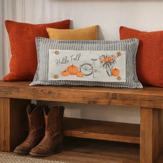 Decorative pillow with 'Hello Fall' text and pumpkins on a wooden bench with orange and red pillows.