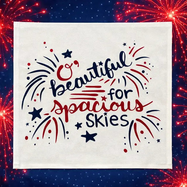 A square white fabric towel or napkin featuring the phrase 'Beautiful for Spacious Skies' in red and navy blue script, surrounded by fireworks and star illustrations. The product is displayed against a festive background of red fireworks and a dark blue starry sky.