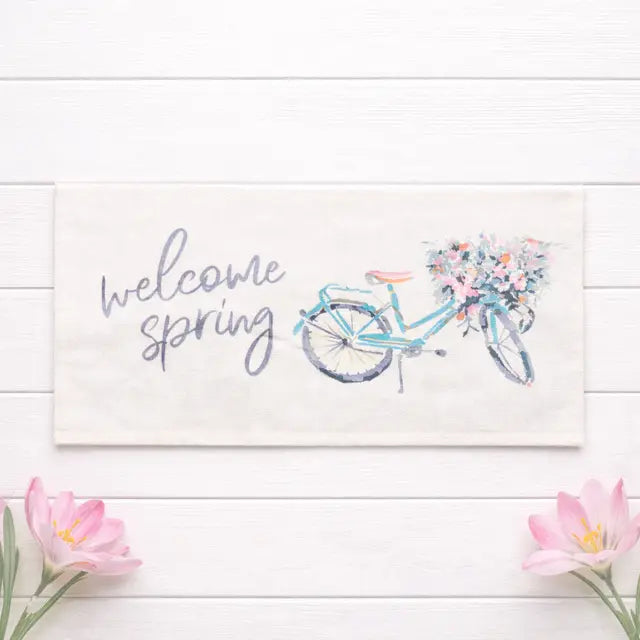 A rectangular cream fabric sign or mat with a watercolor-style illustration of a light blue bicycle carrying pink and purple flowers, next to the words 'welcome spring' in gray script. The item is photographed on a white wood plank background with pink tulips placed in the lower corners.