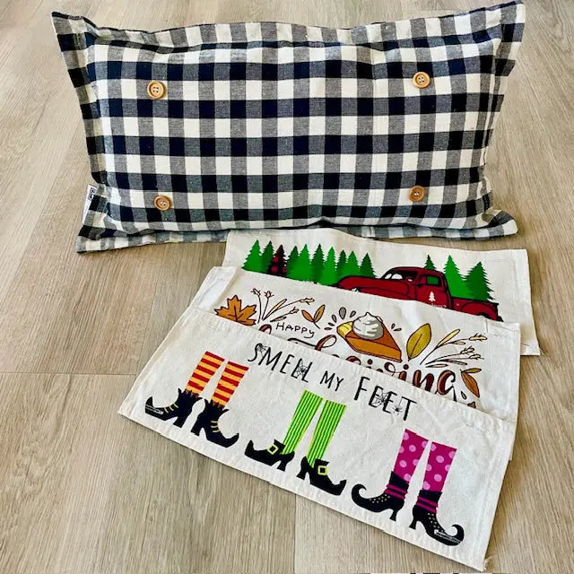 A navy and cream gingham throw pillow with wooden buttons is displayed on a light wood floor next to two embroidered white doilies featuring a fall harvest theme with witch boots and the text 'SMELL MY FEET'.