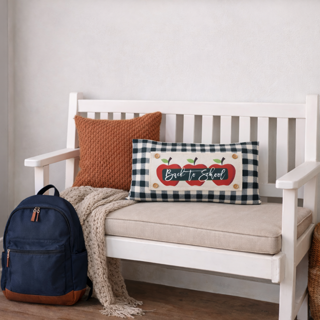 A navy backpack and a beige throw blanket rest beside a white wooden bench, with a decorative rectangular pillow in front. The pillow has a black-and-white gingham border and a central red apple graphic with the words “Back to School” in white script.