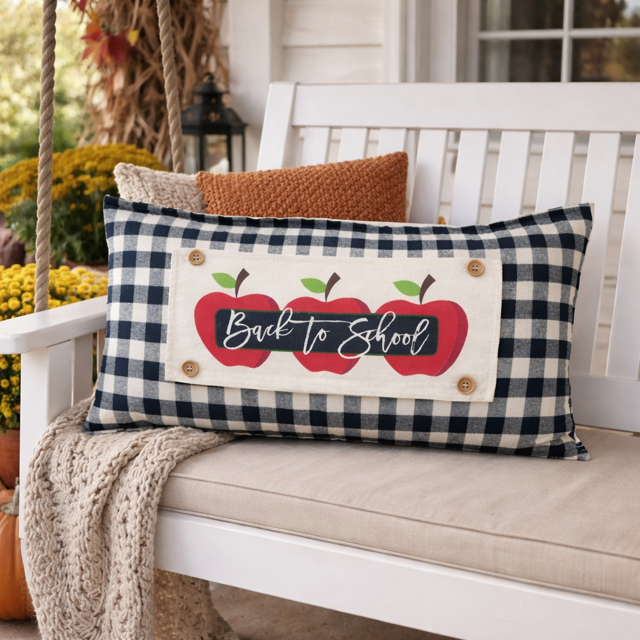 A rectangular decorative throw pillow with a navy and beige gingham pattern, featuring a central cream patch with three red apples and the words 'Back to School' in script. The pillow is styled on a white porch swing with a knit blanket and another pillow in the background.