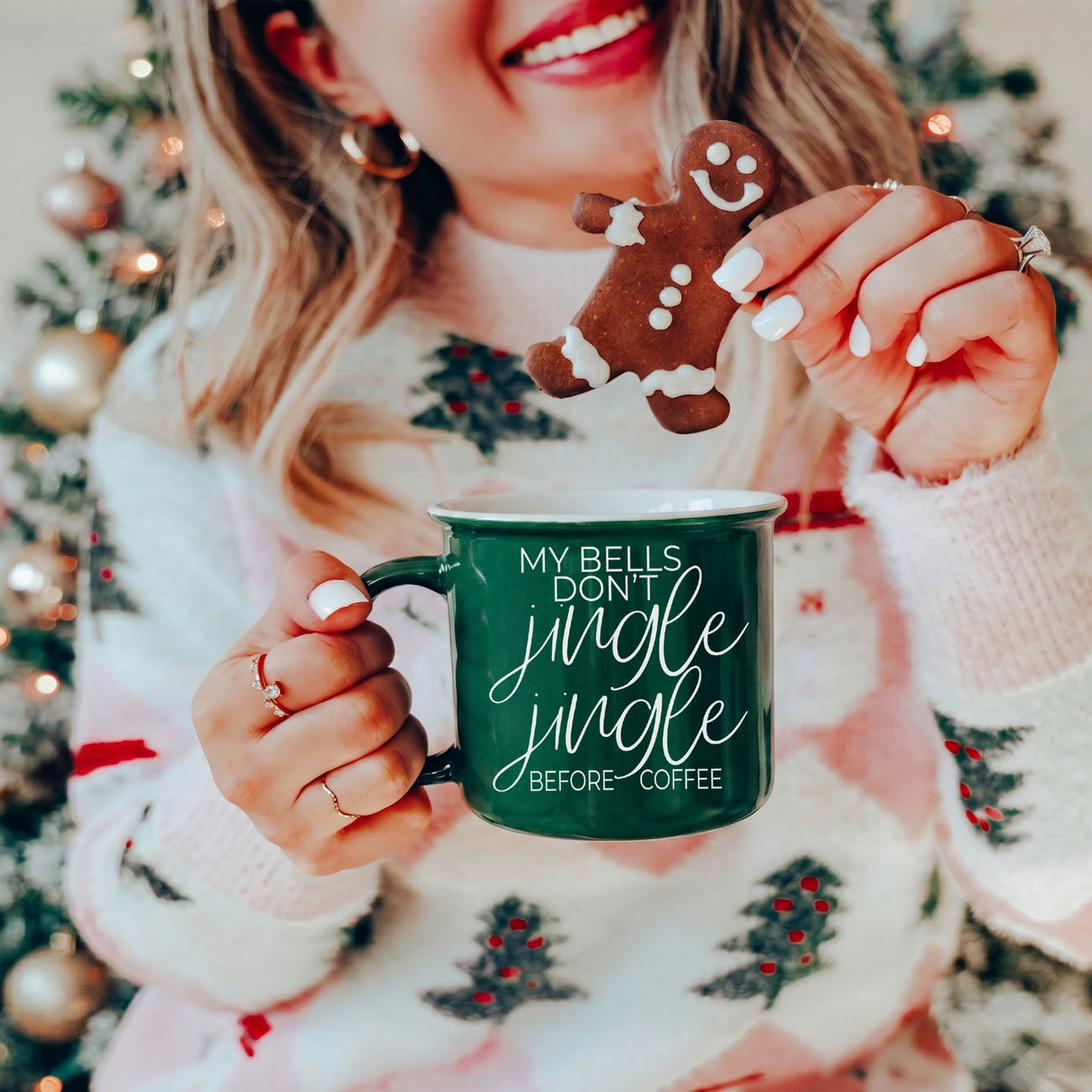Person holding a green mug with a Christmas-themed design and a gingerbread cookie in front of a decorated Christmas tree.