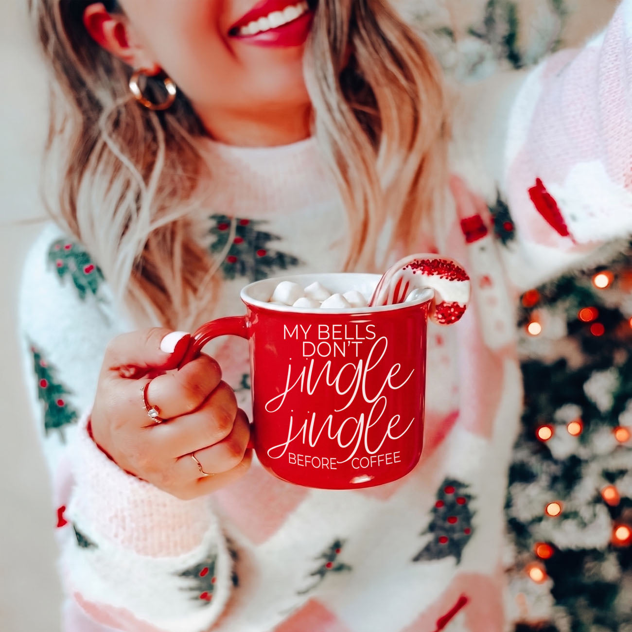 Person wearing a festive sweater holding a red mug with text, surrounded by Christmas lights.