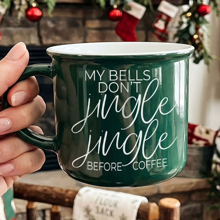 Green mug with 'My bells don't jingle jingle before coffee' text held by a hand, Christmas stockings in the background