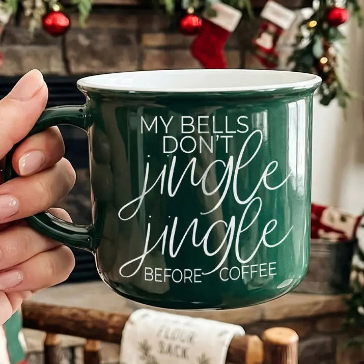 Green mug with 'My bells don't jingle jingle before coffee' text held by a hand, Christmas stockings in the background