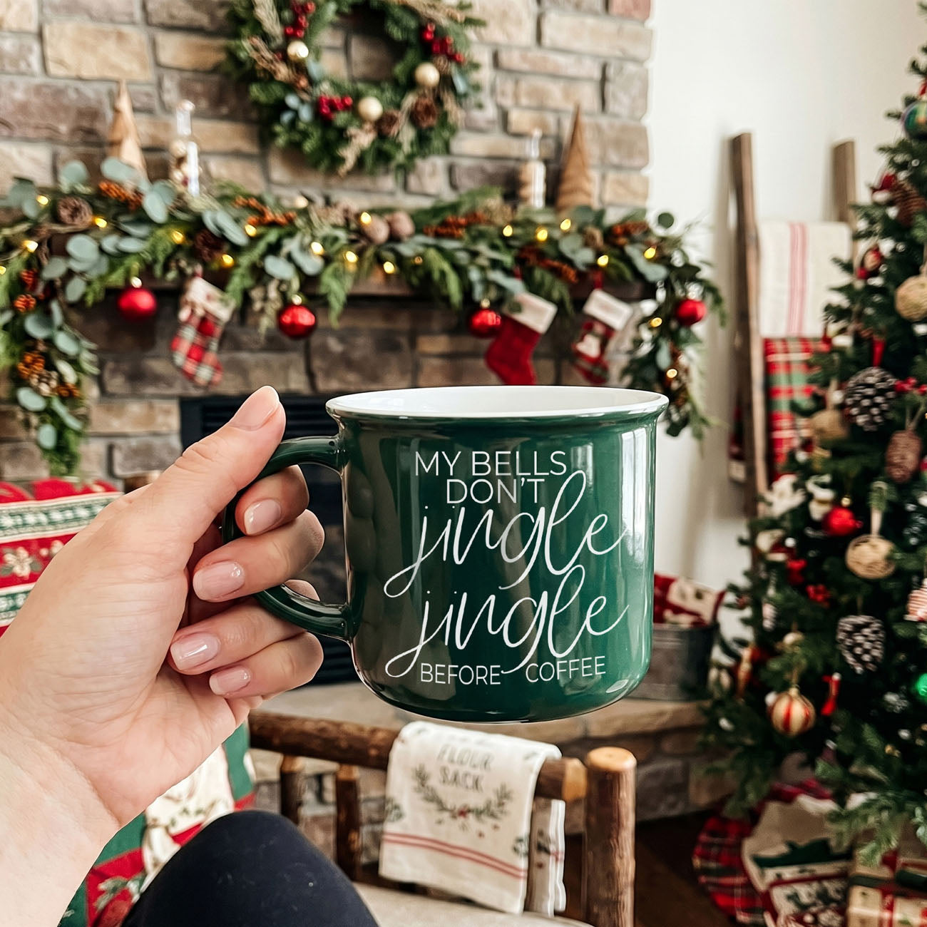 Person holding a green mug with Christmas-themed text in a festive room with decorations.