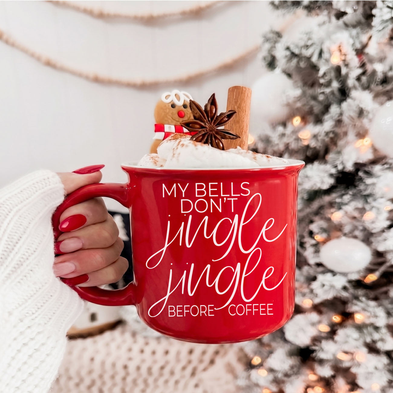 Red mug with festive text held by a hand, decorated with a gingerbread man and cinnamon stick, against a Christmas tree background.