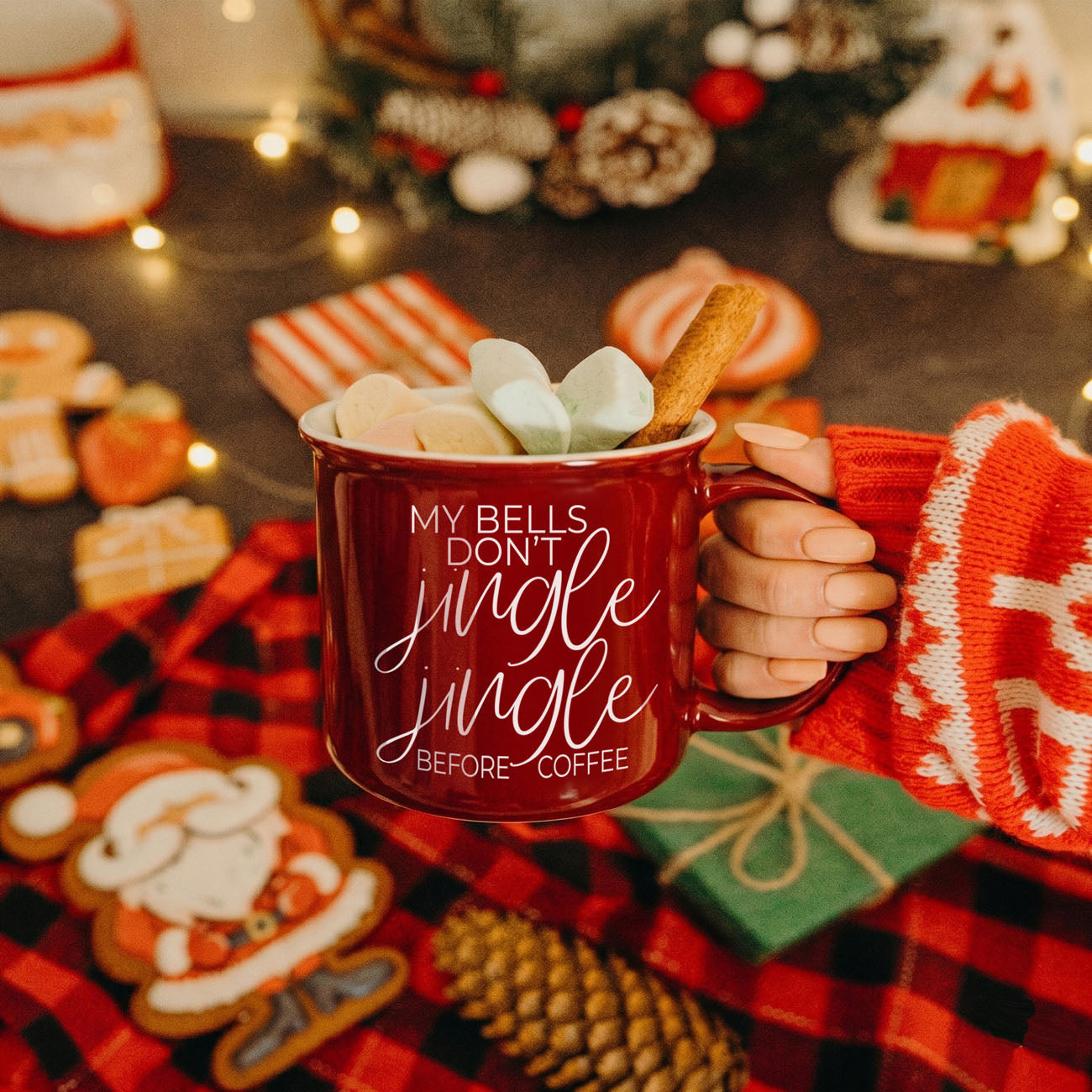 Red mug with text held by a person in a festive setting with cookies and lights.