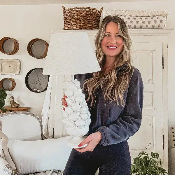 Woman holding white textured lamp in cozy farmhouse living room with rustic decor and wicker basket