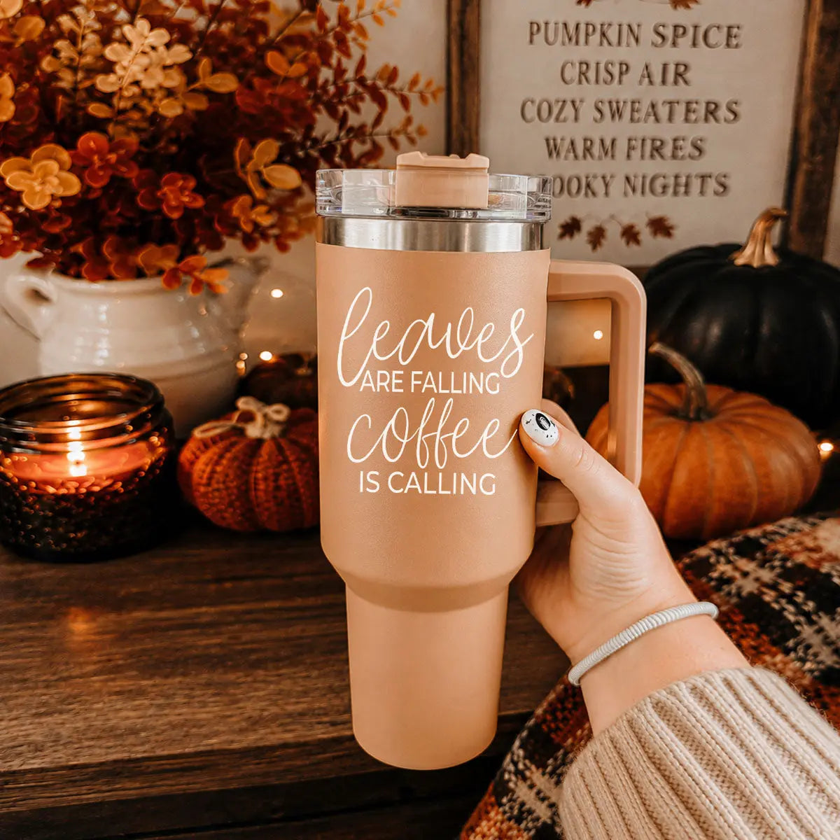 Person holding a 'Leaves are falling, coffee is calling' travel mug with autumn decor in the background.