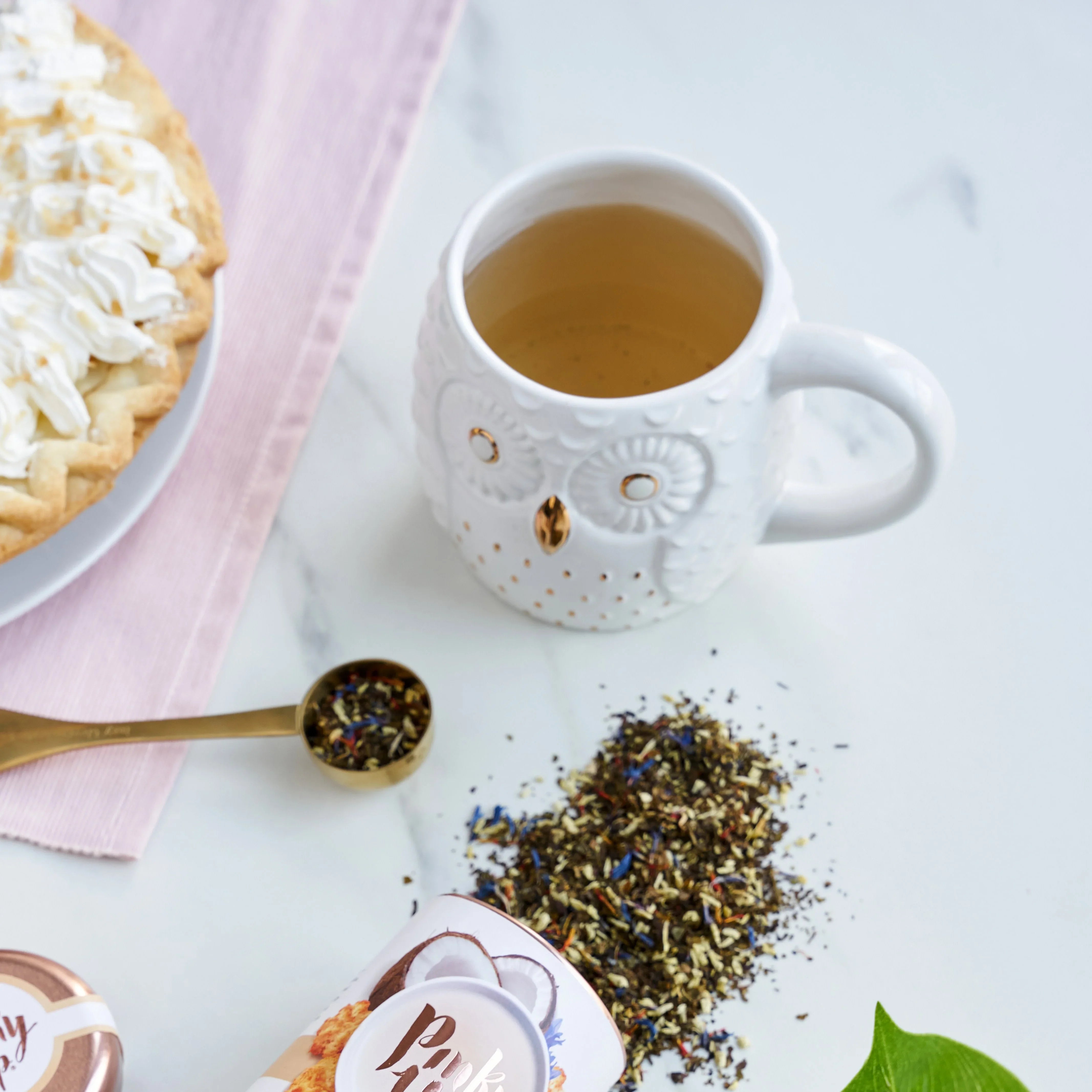 White mug with tea, a plate of cake, and loose tea leaves on a light surface.