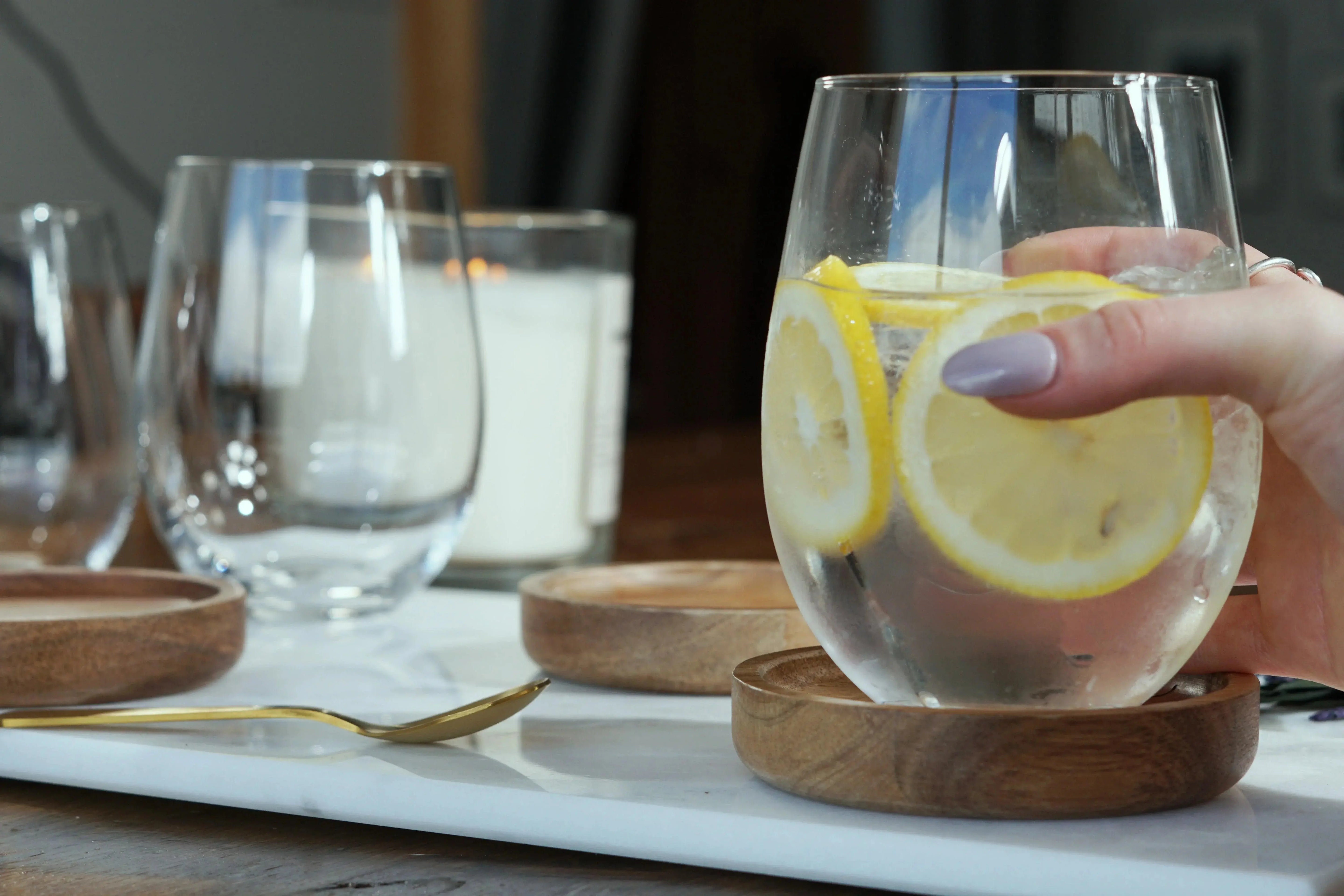 Hand holding stemless wine glass with lemon slices, ice water, on wooden coaster, farmhouse decor style