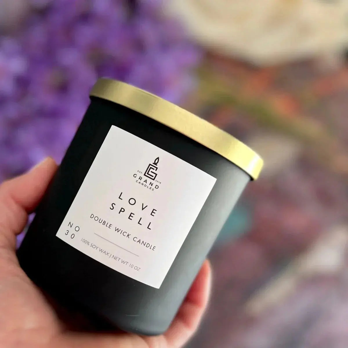 Love Spell double wick soy candle in matte black jar with gold lid, held in hand against purple floral background